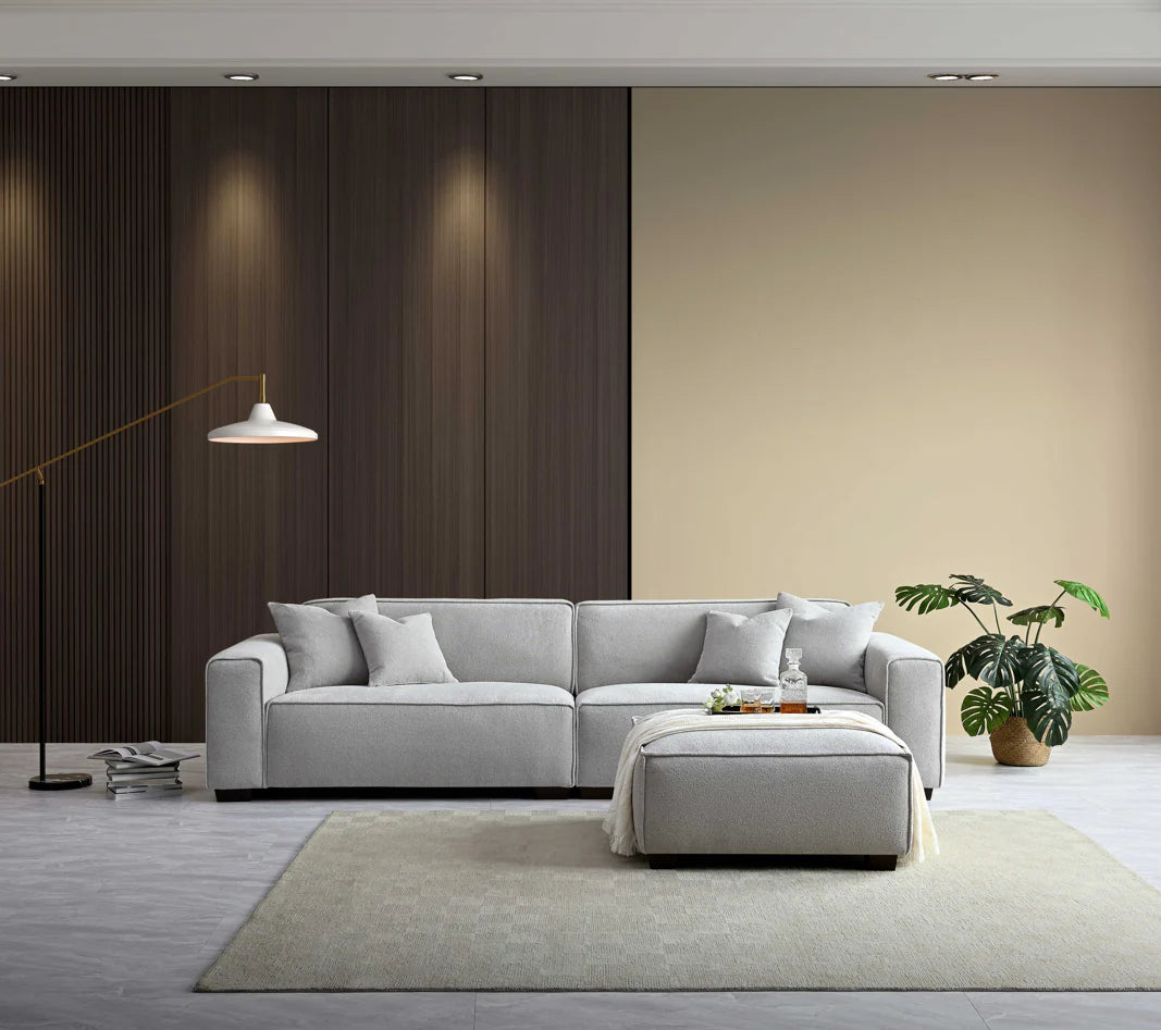 Overhanging spotlight showing 2 seater light grey sofa with footstool in the foreground. Beige background wall with wood detail. 
