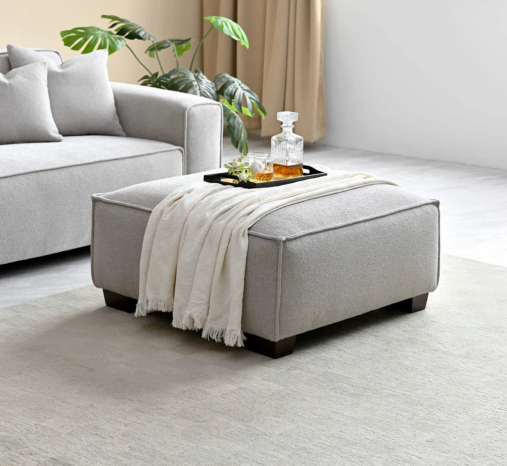 Footstool complete with beige blanket and whiskey tray, light grey sofa in background with plant. 