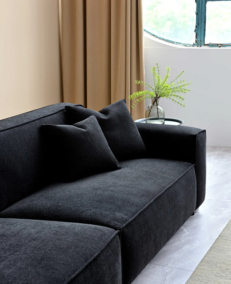 Close up of soft touch grey sofa with 2 cushions and a side table, hanging curtains in the background and a circular window. 