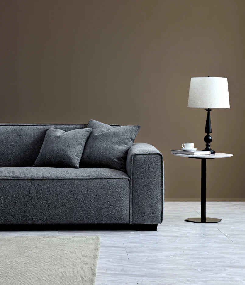 Off centre shot of 2 seater grey sofa with side table and white lampshade in a plain beige background. 